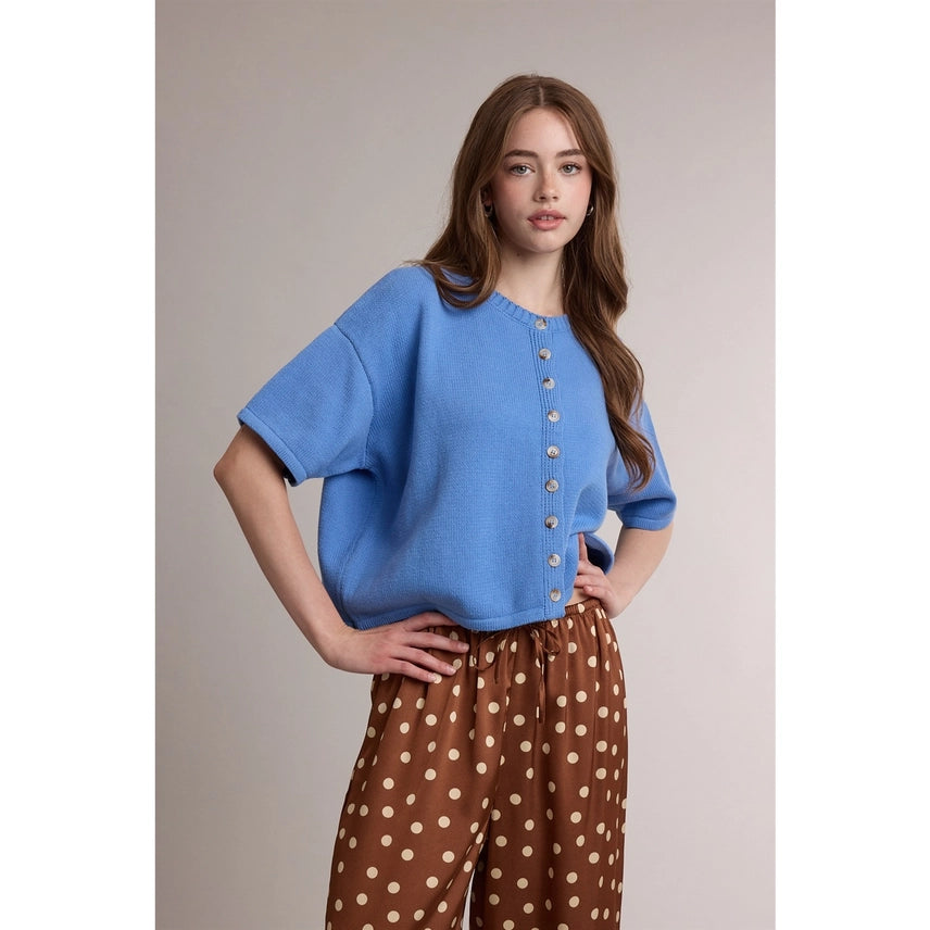 A woman modeling a light blue short-sleeve knit top with a button-down front, paired with brown wide-leg pants featuring a beige polka dot pattern, set against a plain beige studio background.