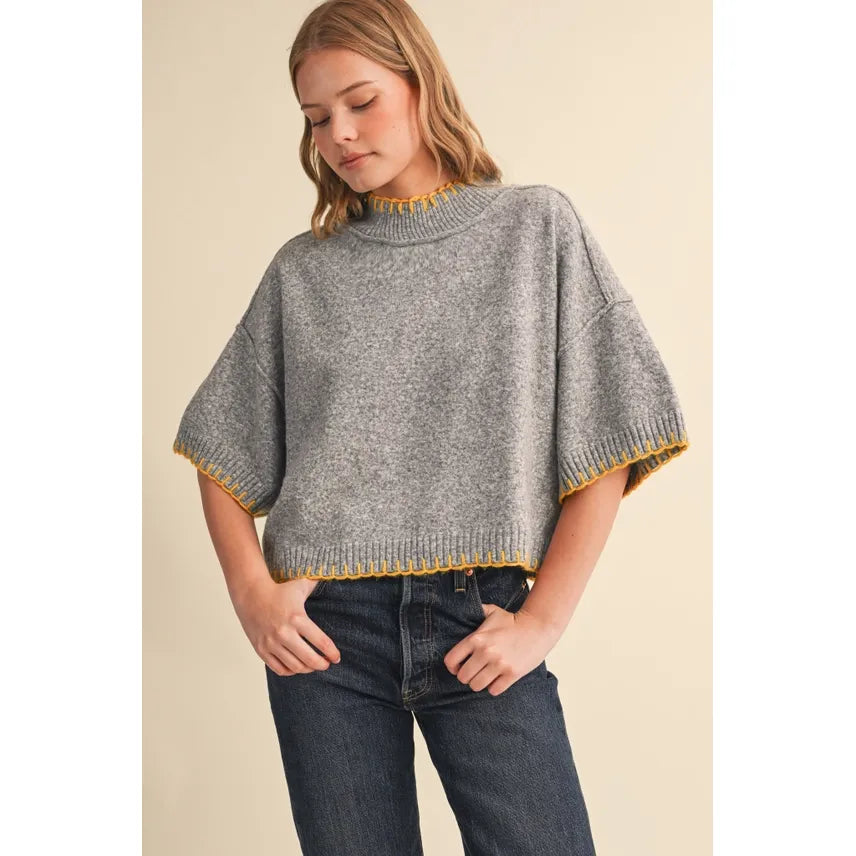 A woman wearing a gray knit cropped sweater with yellow trim on the collar and sleeve cuffs, paired with dark blue jeans, against a solid beige background.