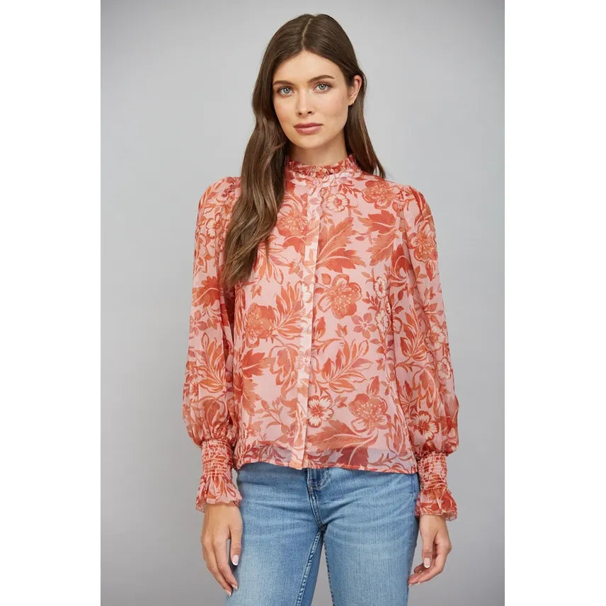 A woman modeling a long-sleeve high-neck blouse with a pink and orange floral pattern, paired with blue jeans, against a solid gray studio background.