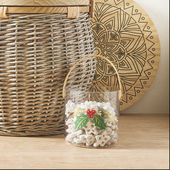 A wicker gift basket set with a large central basket holding a bottle and two glasses, surrounded by matching wicker accessories, a small clear bucket, a round tray, and a small jar on a tabletop against a plain light background.