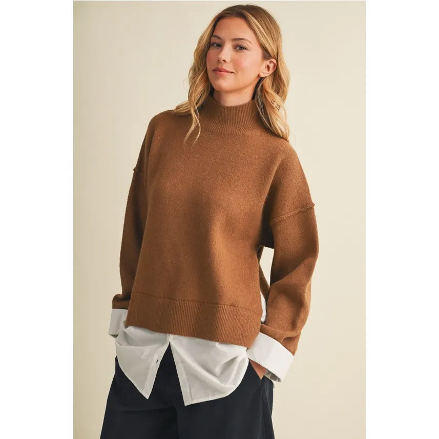 A woman wearing an oversized camel brown knit mock neck sweater, layered over a white button-down shirt and dark trousers, set against a plain beige background.