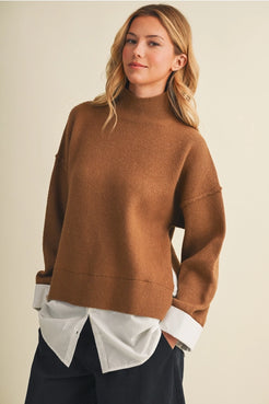 A woman wearing an oversized camel brown knit mock neck sweater, layered over a white button-down shirt and dark trousers, set against a plain beige background.