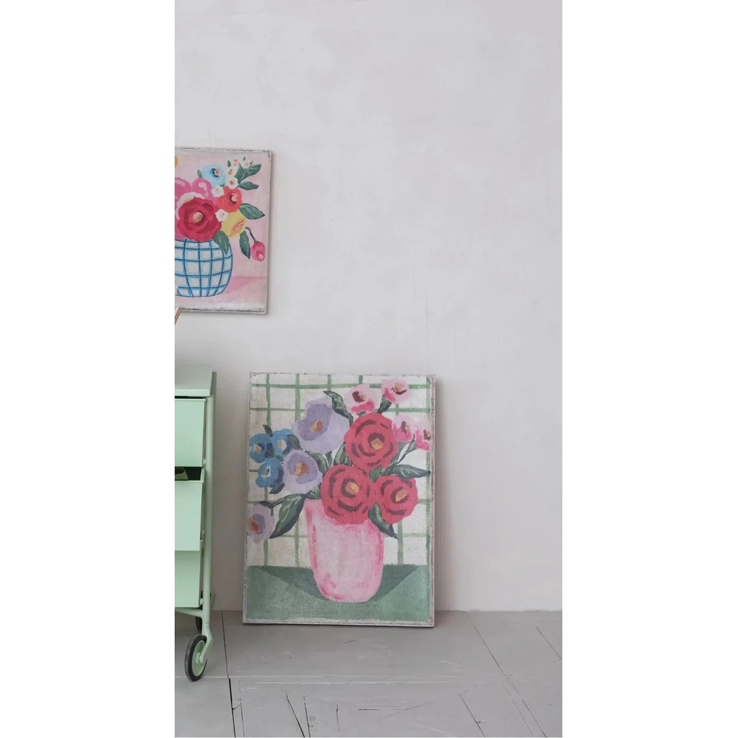 Light green side table with a lamp and books, next to floral artwork on a white wall.