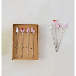 Three small open kraft-paper trays holding decorative metal picks or pins with colorful tops, arranged on a light textured wall. A translucent green glass cup holds two picks in the center.